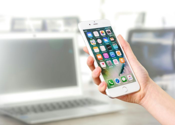 Close-up view of a smartphone displaying apps, held by a hand, with a blurred laptop in the background.